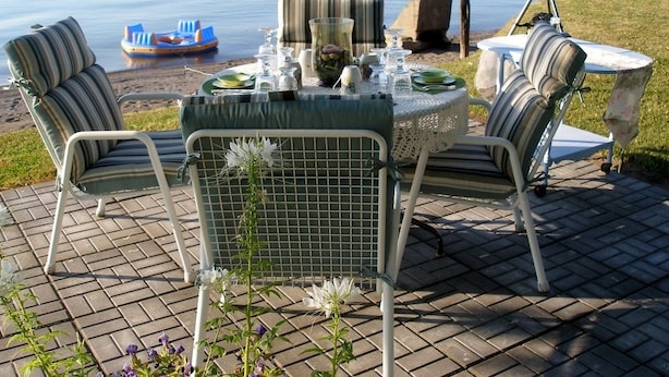A patio with a table set for dining, surrounded by chairs, overlooking a calm body of water.