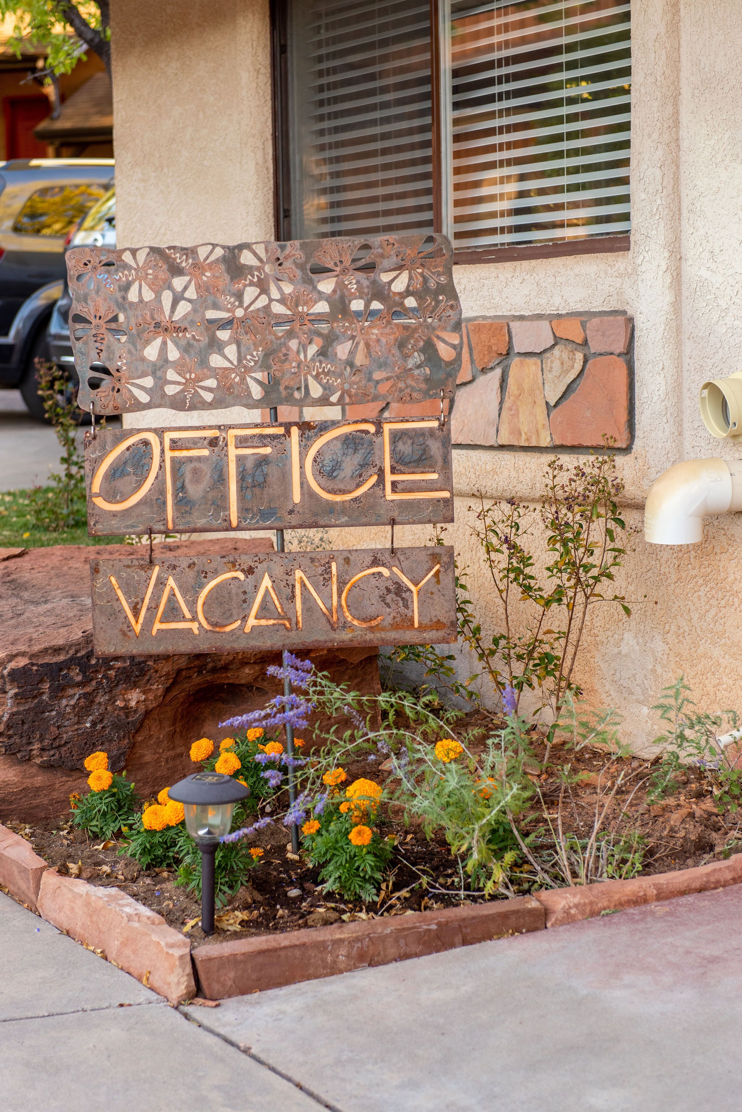 A wooden sign with "OFFICE VACANCY" written on it, placed in front of a building with a window and a planter with flowers.