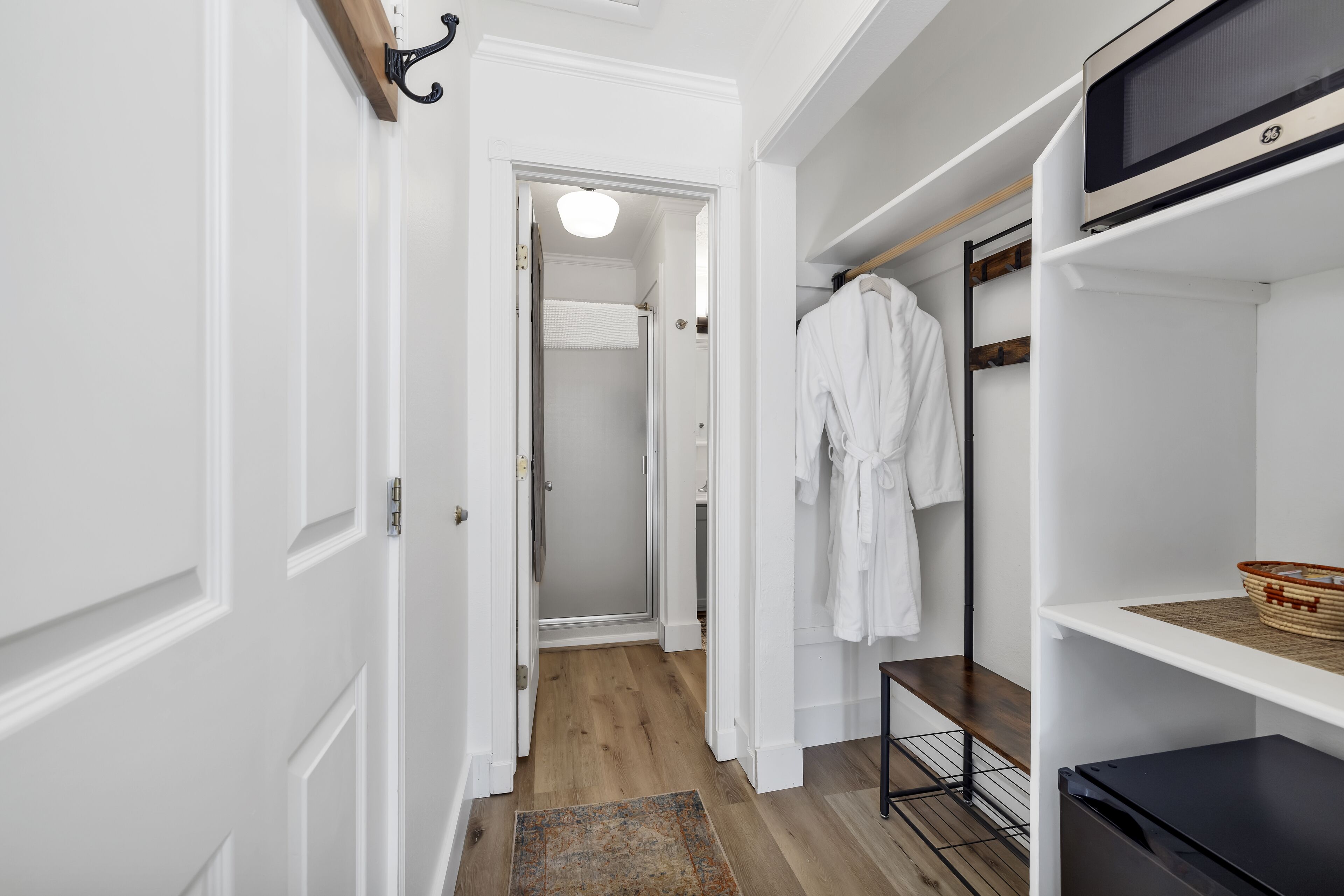 A hallway with a white door, a wooden floor, a white robe hanging on a rack, and a black appliance on the right.