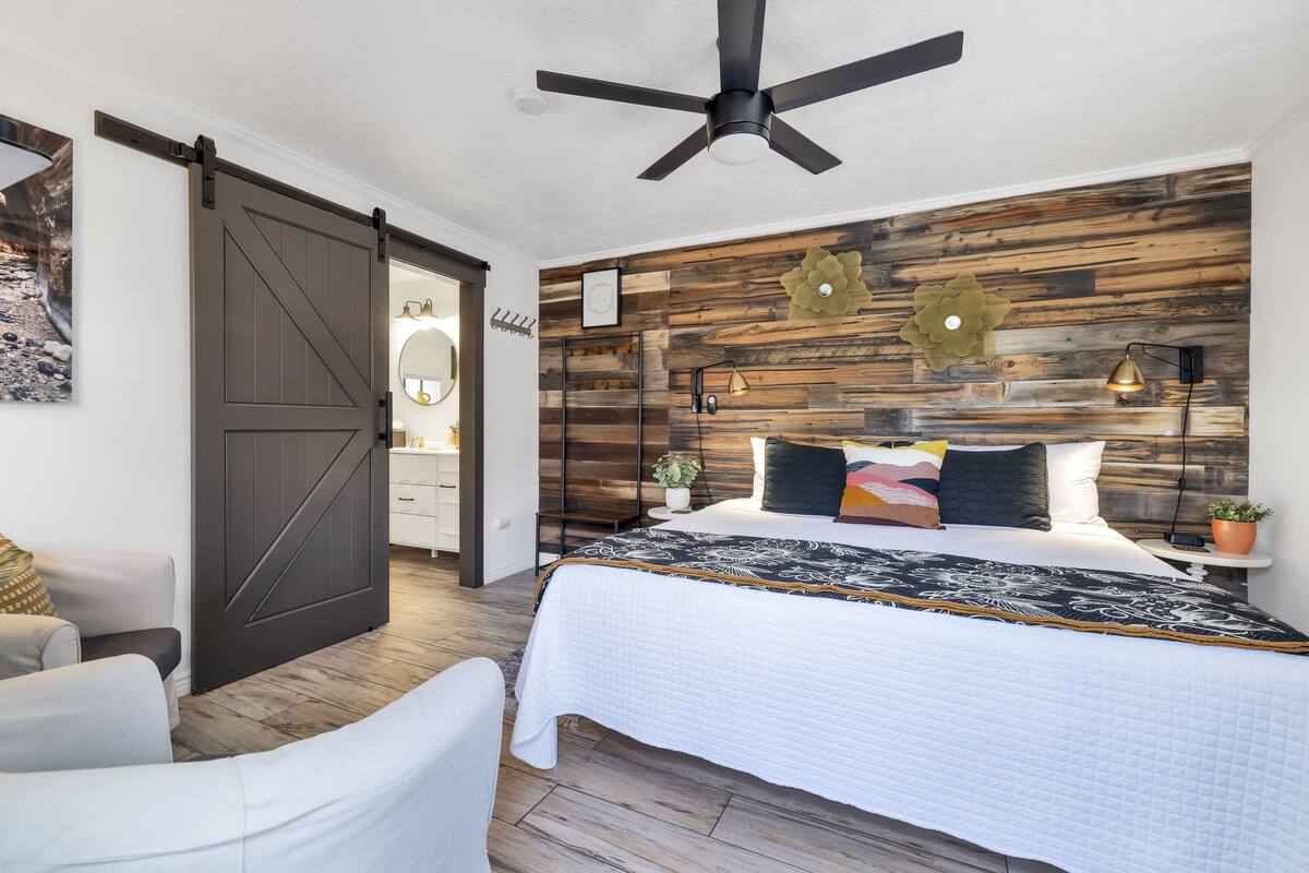 A bedroom with a bed, a ceiling fan, a wooden headboard, a nightstand, and a wall-mounted lamp.