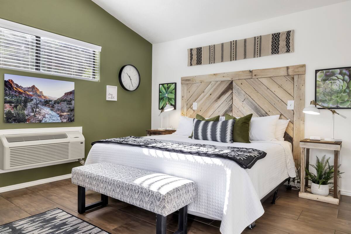 A bedroom with a bed, a bench, a wall-mounted clock, a wall-mounted air conditioner, and a window with blinds.