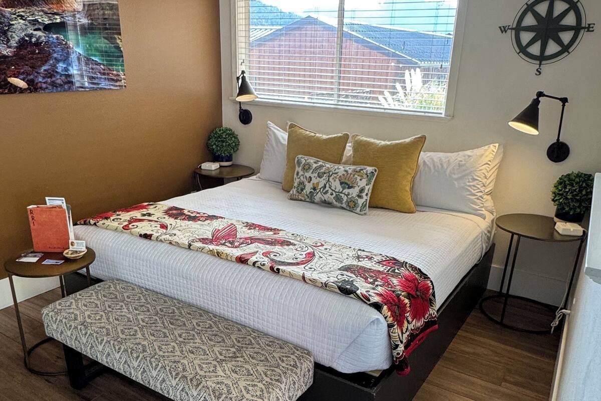 A bedroom with a bed, bedside tables, a bench, a window with blinds, a compass wall decoration, and a painting on the wall.
