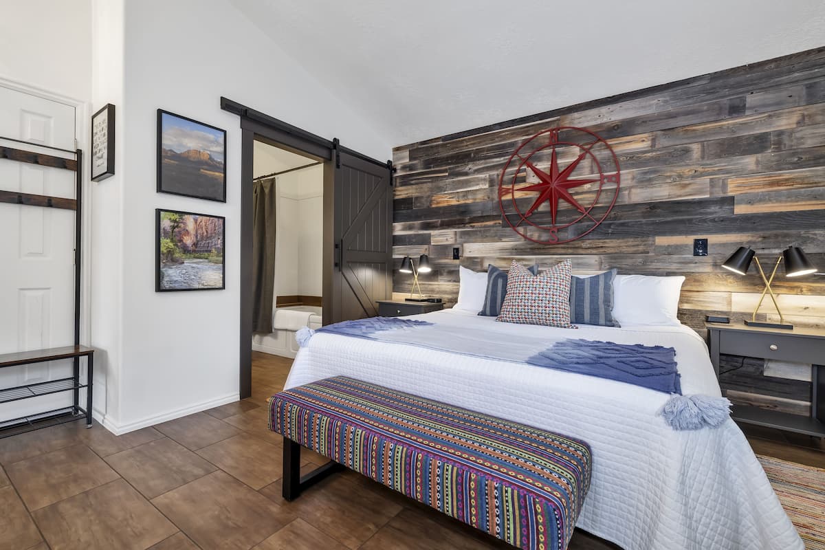 A bedroom with a large bed, a colorful bench, a wooden wall with a compass, and a bench with a lamp.