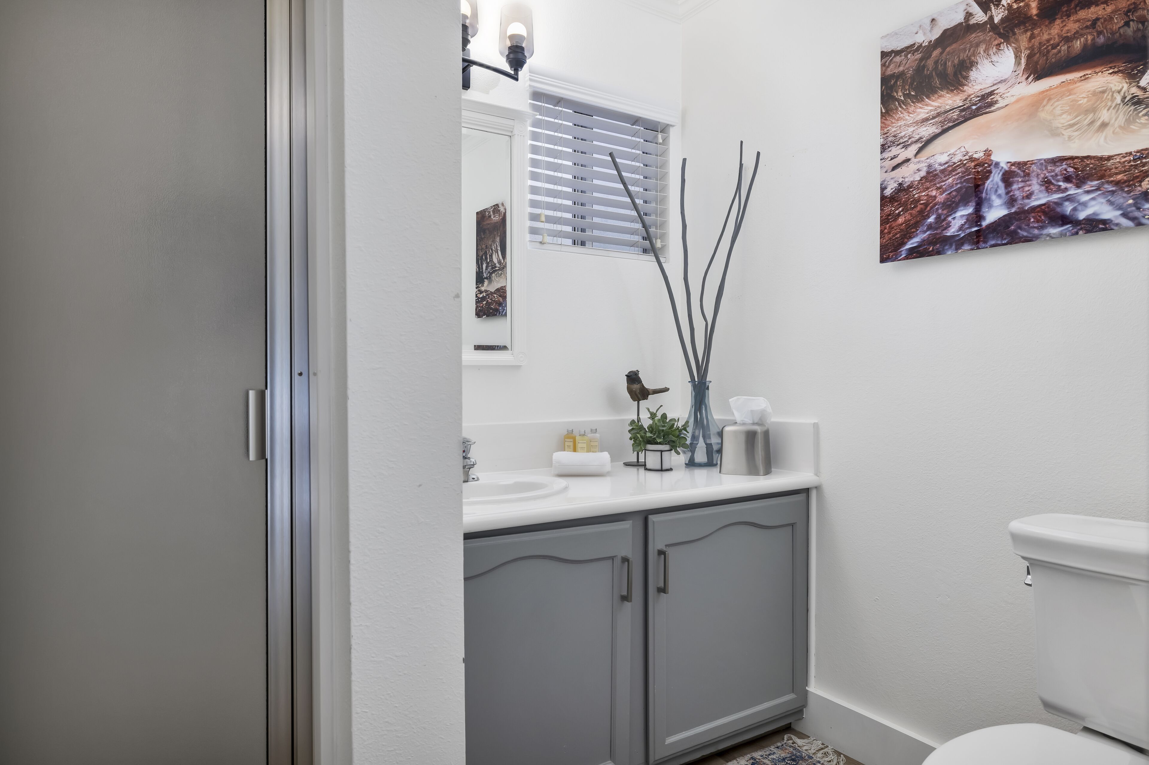A bathroom with a white sink, grey cabinets, a toilet, a window with blinds, and a painting on the wall.