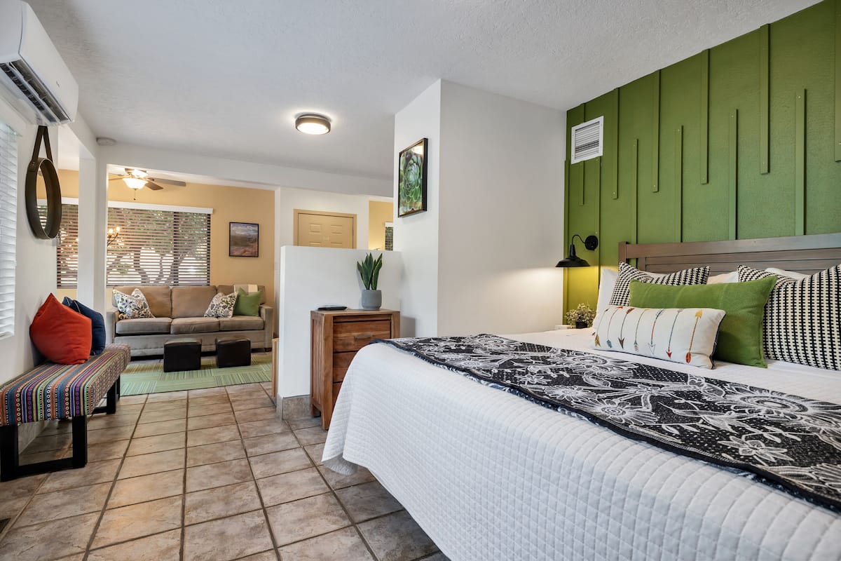 A bedroom with a bed, a green headboard, a bedside table, a nightstand, a bench, a sofa, and a window with blinds.