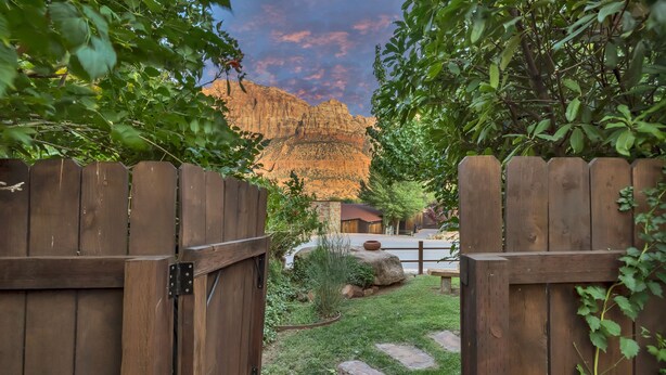 Un portón de madera da acceso a un jardín con un sendero de piedra y abundante vegetación, con una montaña al fondo.