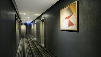 A hotel hallway with dark walls, striped carpet, and a geometric painting on the wall.