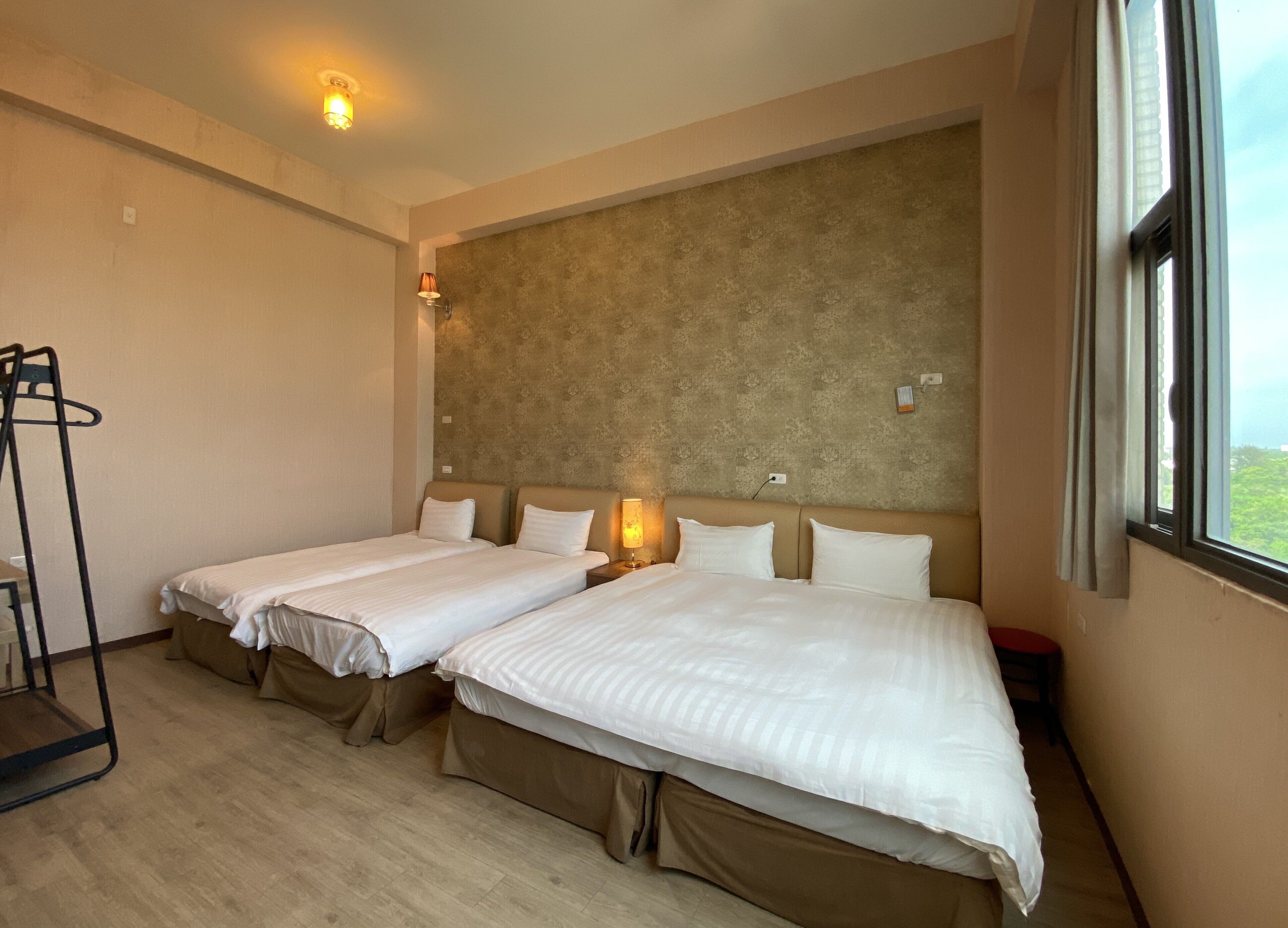 Une chambre d’hôtel avec deux lits, un plancher en bois, une tête de lit agrémentée d’un mur texturé, une fenêtre avec des rideaux et une lampe fixée au mur.