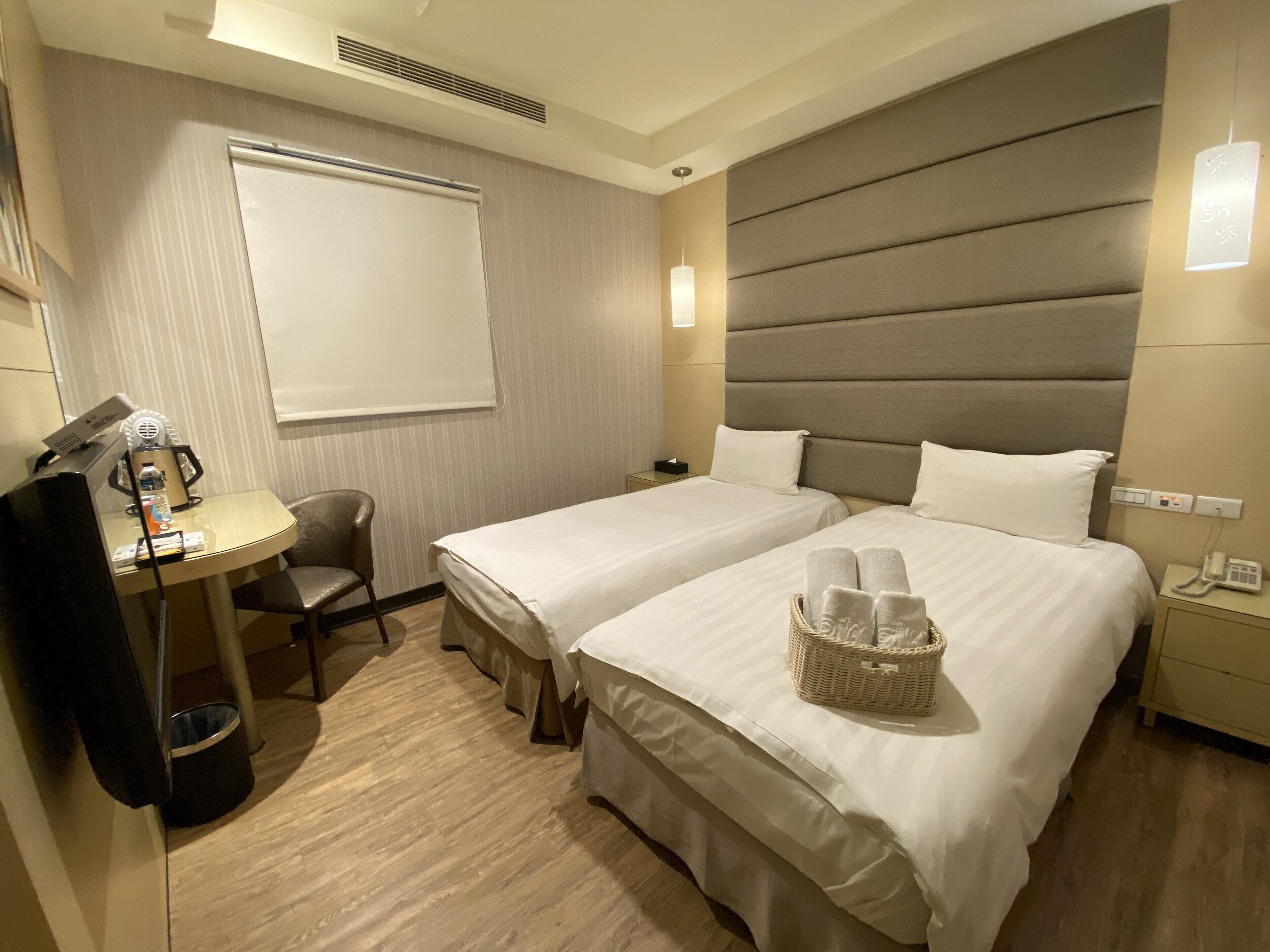 A hotel room with two beds, a desk with a lamp, a chair, a waste bin, and a basket of towels.