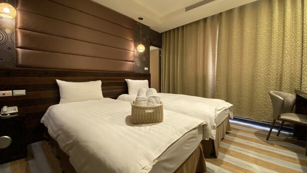 Habitación de hotel con dos camas, cabecera de madera, teléfono en la mesita de noche, cesta de mimbre con toallas y una silla.