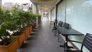 Un espacio de estar al aire libre con techo, mesas y sillas, plantas en macetas y vista a la calle.