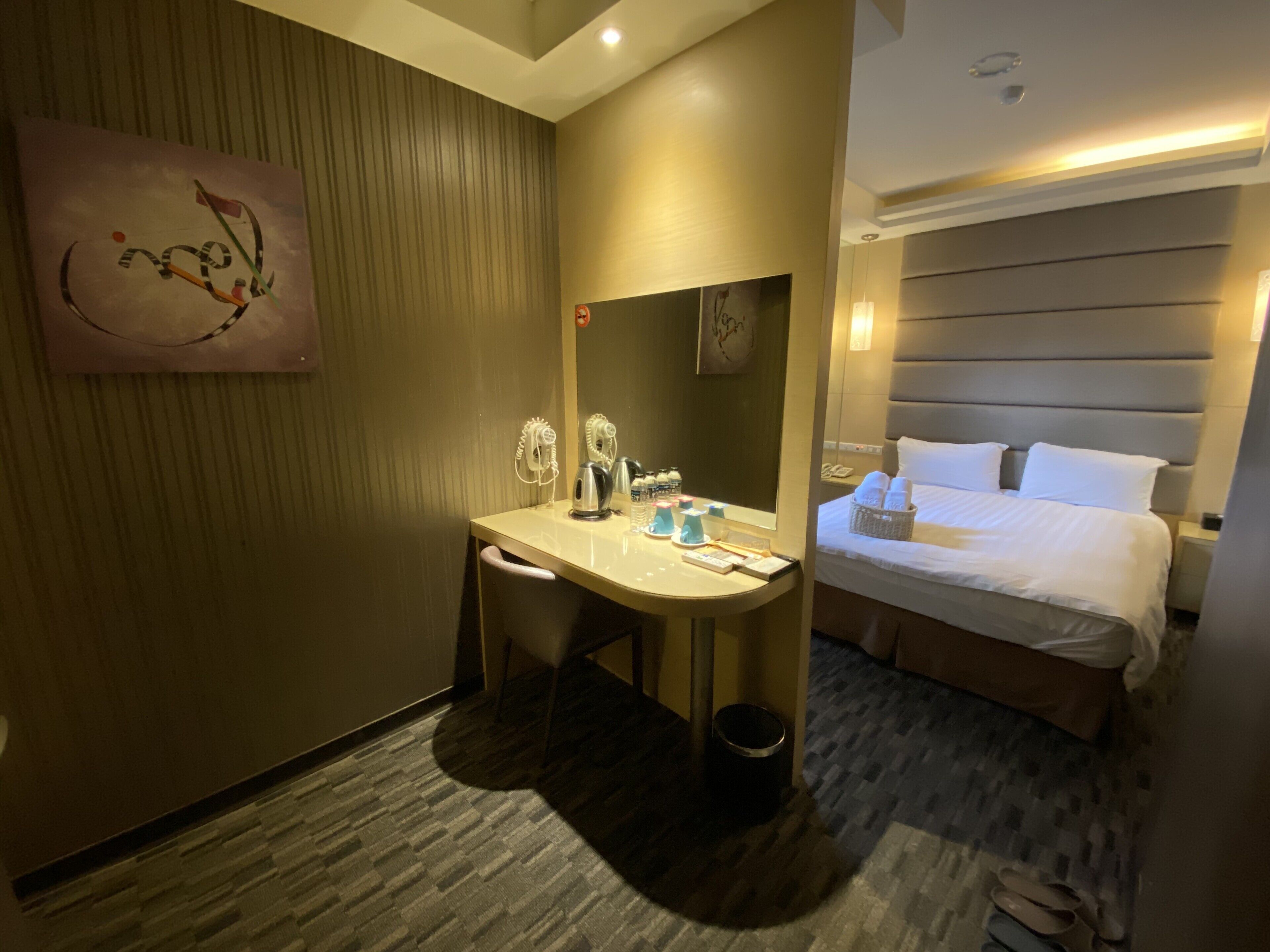 A hotel room with a bed, a desk with a chair, a mirror, a trash bin, and a wall-mounted artwork.