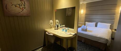 A hotel room with a bed, a desk with a chair, a mirror, a trash bin, and a wall-mounted artwork.