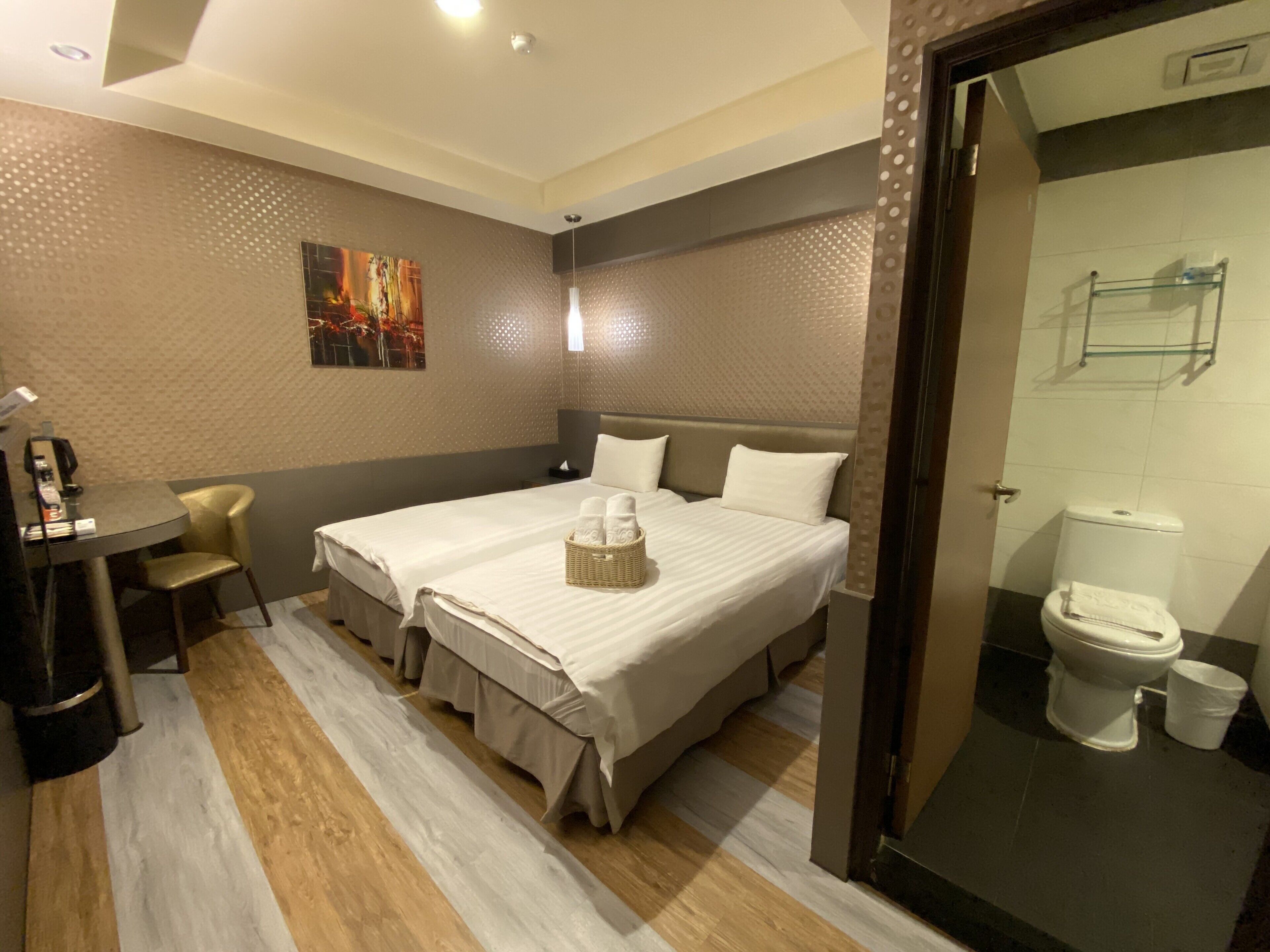 Habitación de hotel con dos camas, un escritorio con silla, un cuadro en la pared y un baño con inodoro y lavamanos.