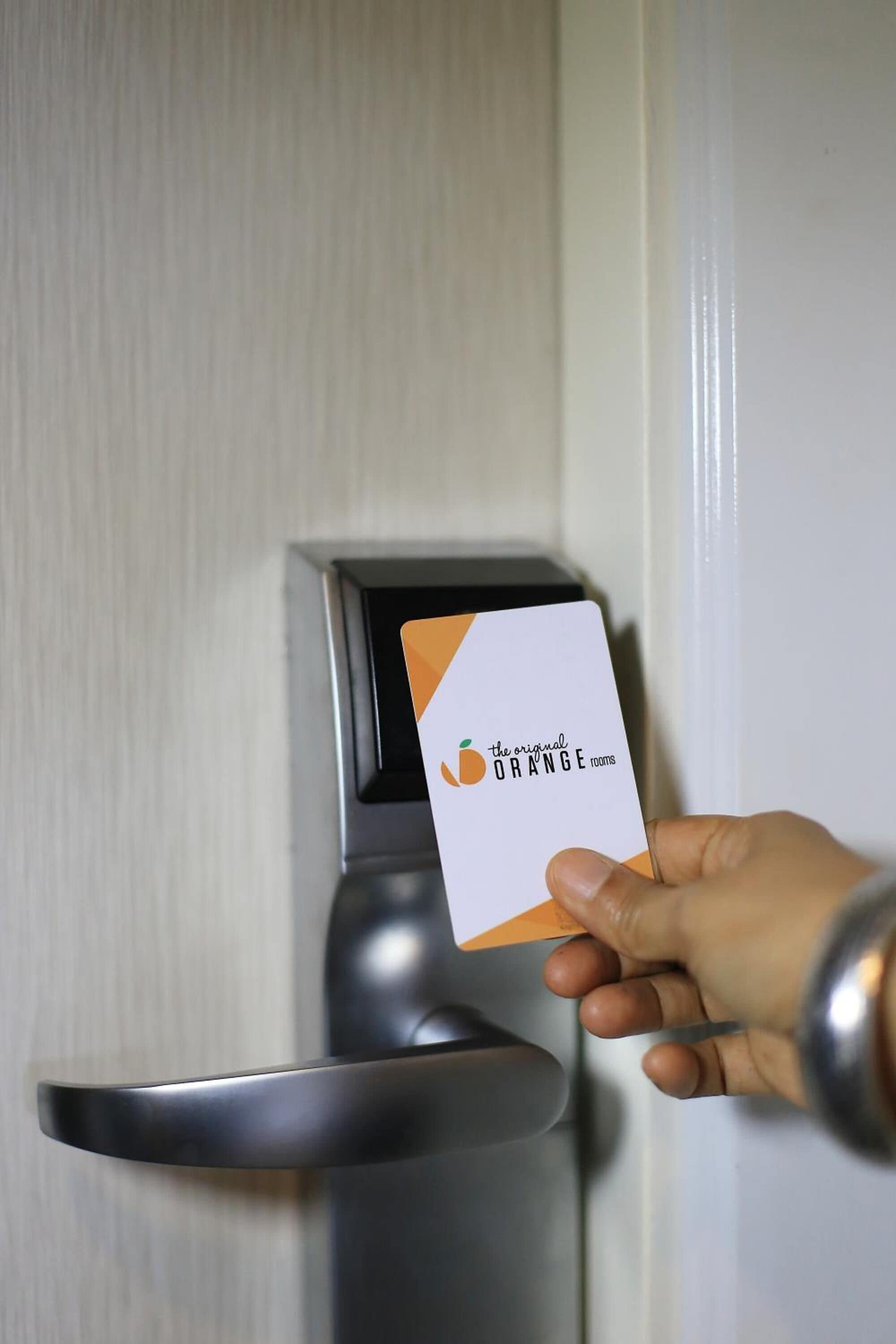 A hand holding a card with the text "the original ORANGE rooms" in front of a door with a modern lock mechanism.