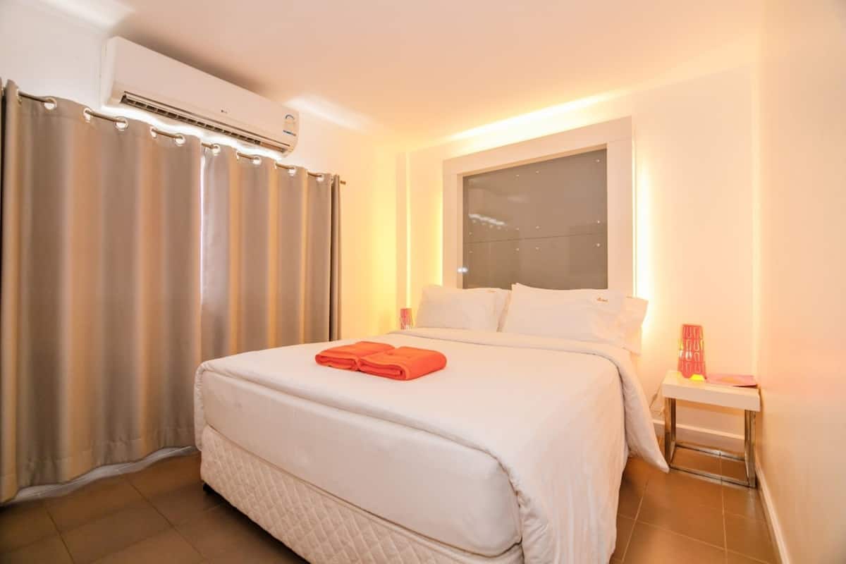 Una habitación de hotel con cama, cortinas, mesita de noche y aire acondicionado.