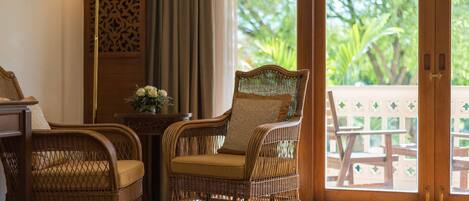 A wicker chair with a cushion, a small table with a vase of flowers, and a view of greenery through a sliding door.