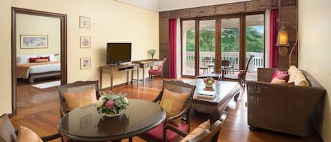 Habitación de hotel con comedor, sofá, televisión y balcón con vistas a árboles.