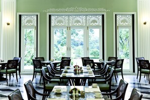 Un comedor con una mesa grande dispuesta para una comida, rodeada de sillas, y vista a un follaje exuberante a través de las ventanas.