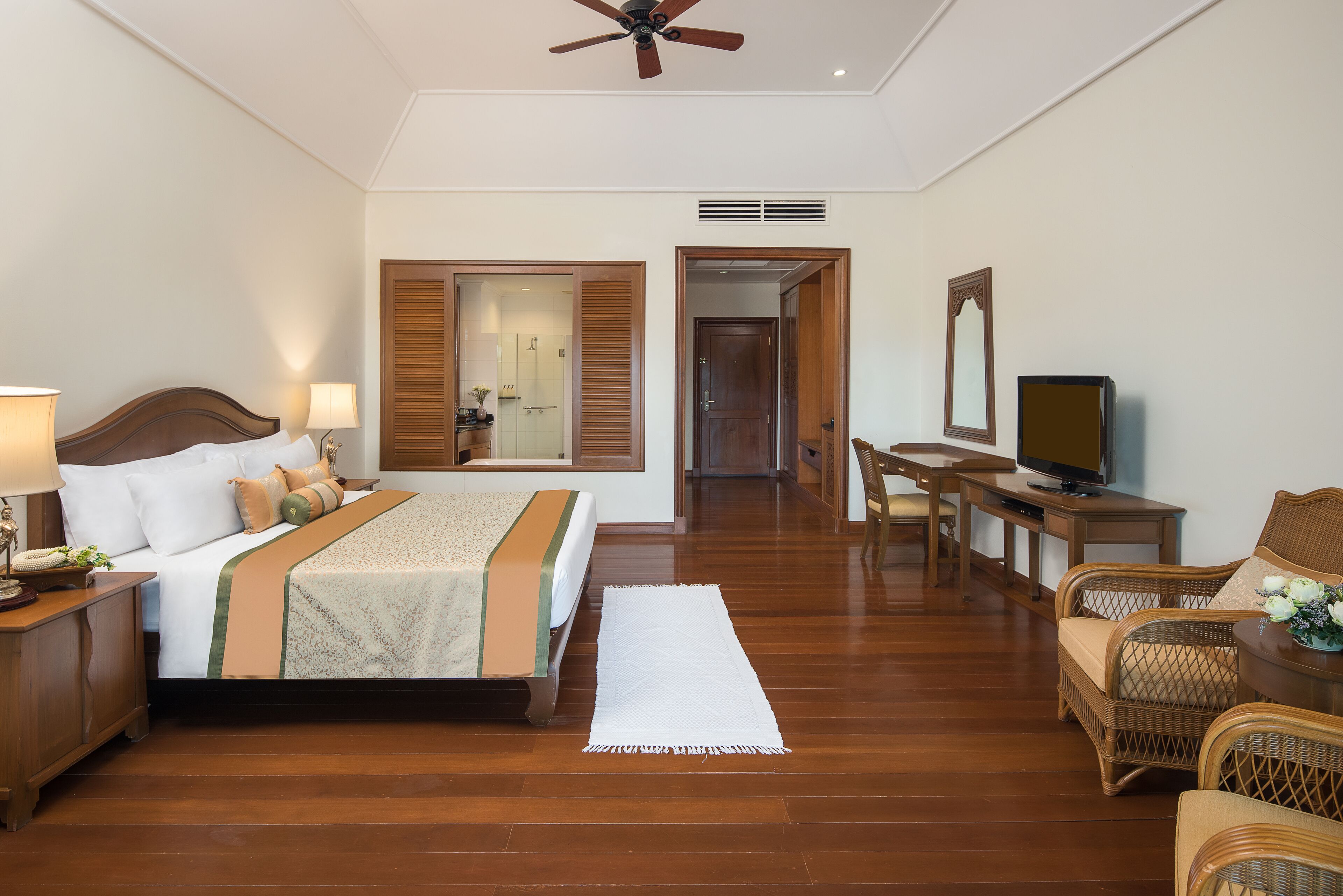 A hotel room with a bed, bedside table, lamp, TV, and wicker furniture.