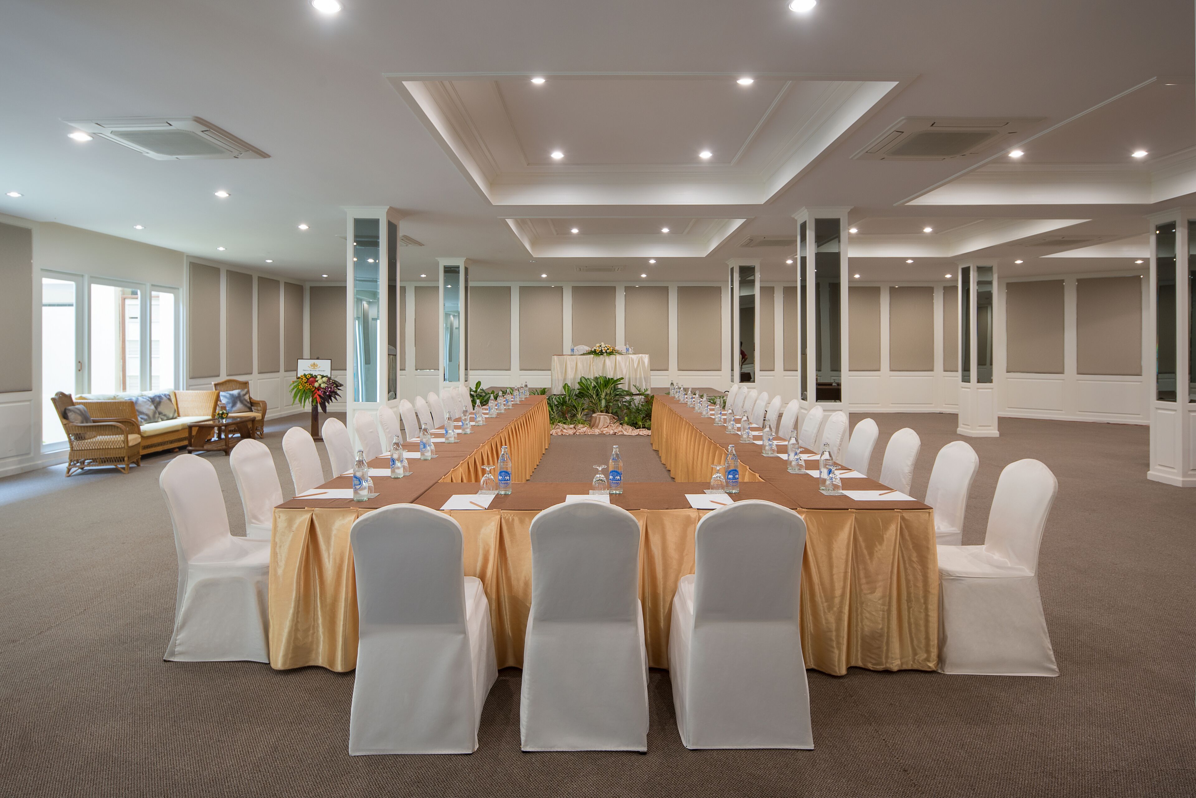 Uma sala de conferências com uma mesa comprida, cadeiras brancas e um tapete bege.