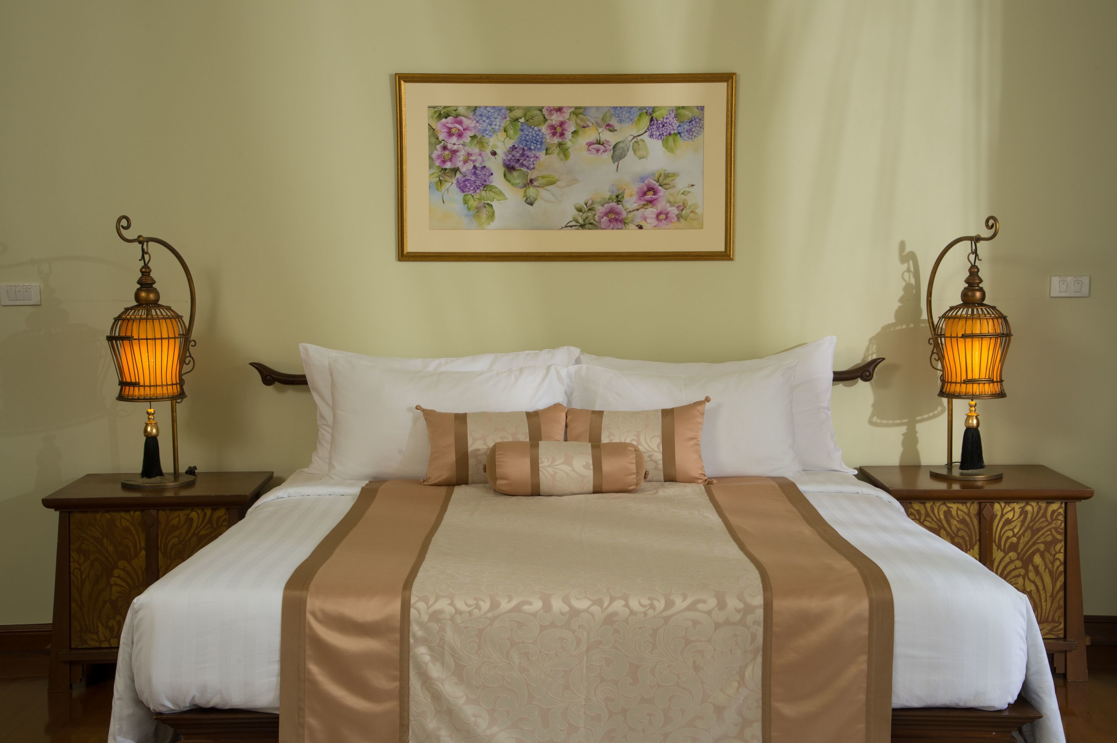 A neatly made bed with white and brown bedding, two bedside lamps, and a floral painting on the wall.