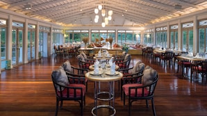 A spacious dining area with multiple round tables and chairs, large windows, and a high ceiling with exposed beams.