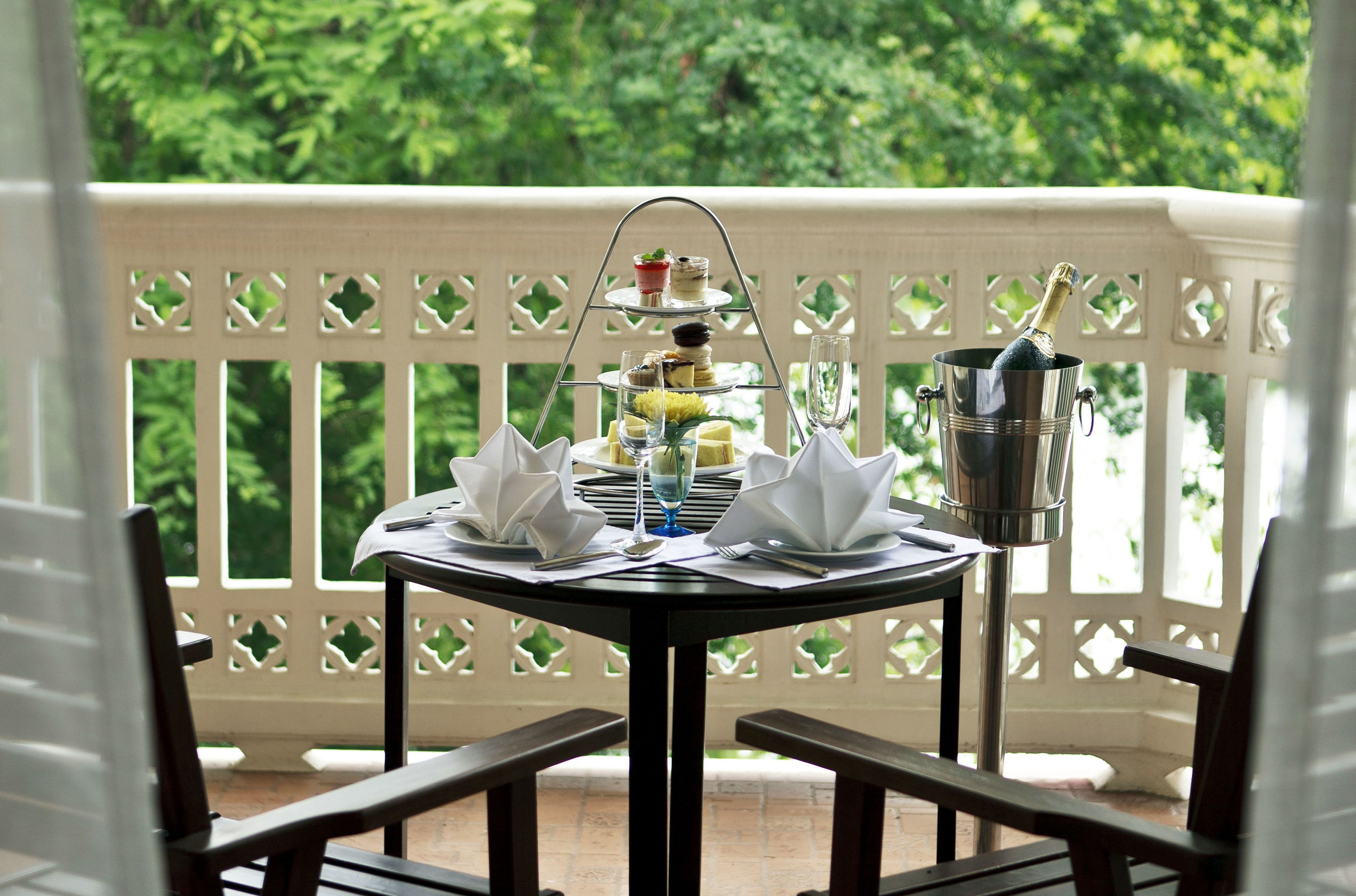 A table set for a meal with a three-tiered dessert stand, a bottle of champagne, and a glass on a balcony with a white lattice railing.