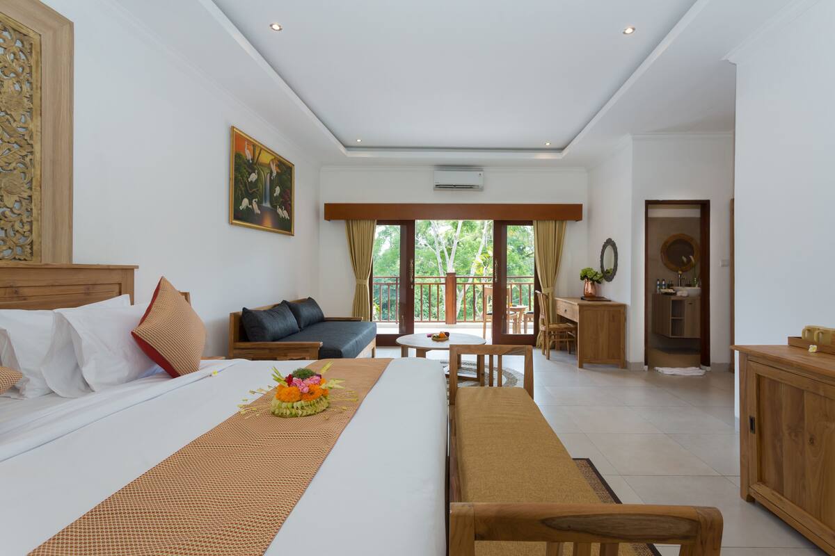 A hotel room with a bed, a sofa, a dining table, and a balcony with a view of trees.