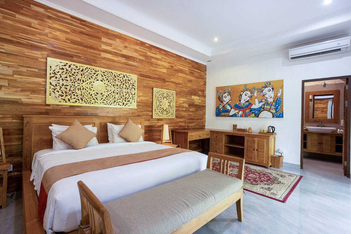 A hotel room with a large bed, wooden headboard, a desk, a bench, a bathroom with a sink and mirror, and a painting on the wall.