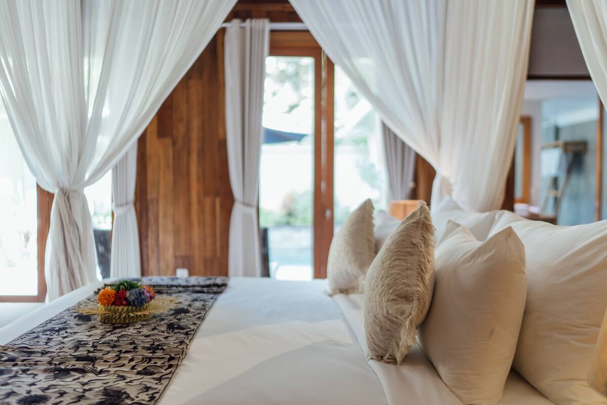 A bedroom with a bed, white curtains, and a view of the outdoors.