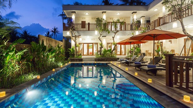 A pool area with lounge chairs and umbrellas, surrounded by lush greenery and a two-story building with balconies. - Puri Payogan Villa Ubud (Ubud)