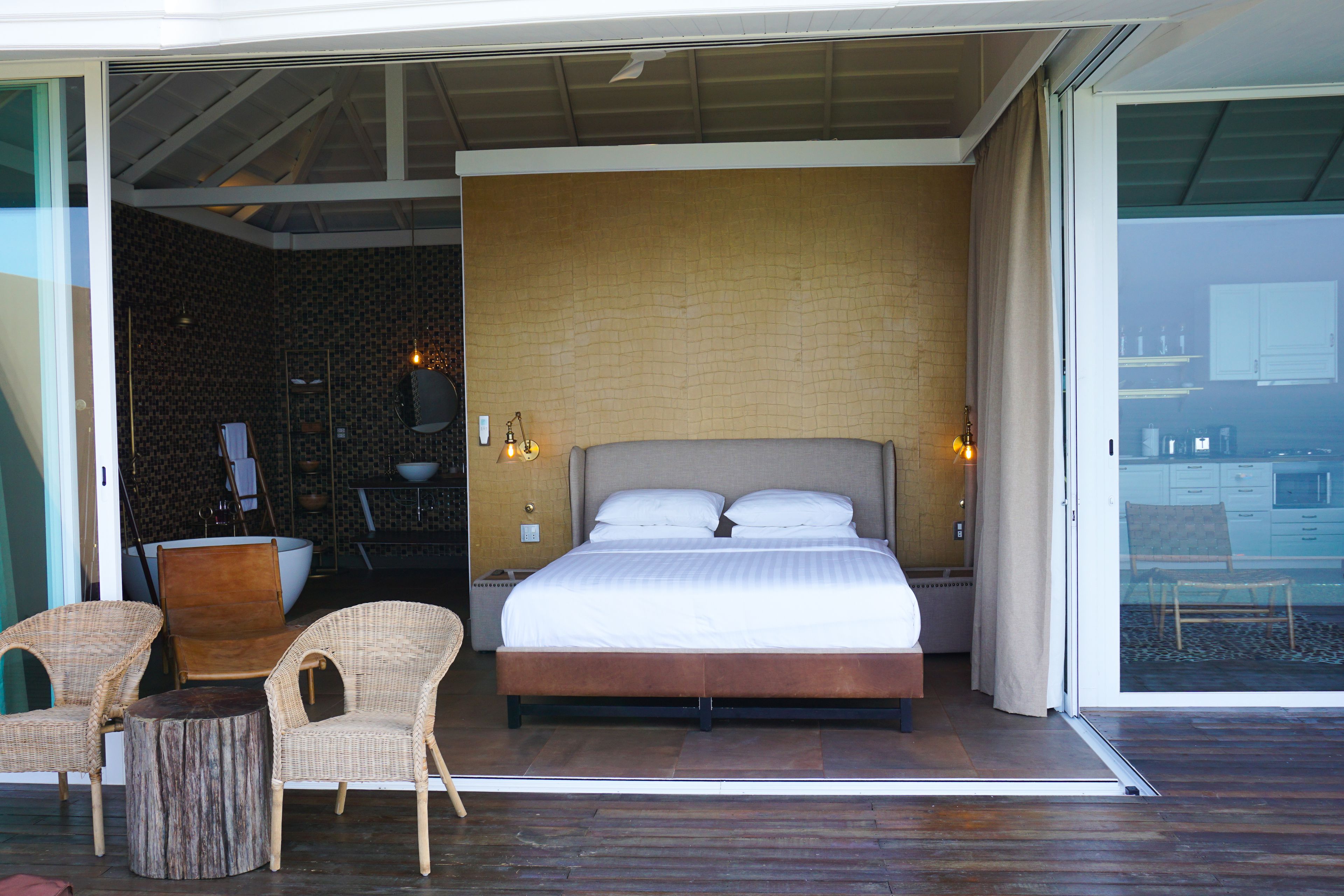 Une chambre moderne avec un grand lit, deux fauteuils en osier, une petite table ronde et une porte vitrée donnant sur un espace extérieur.