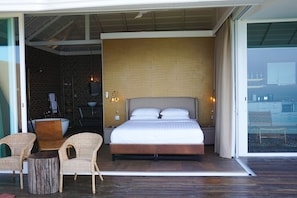 A modern bedroom with a large bed, two wicker chairs, a small round table, and a glass door leading to an outdoor area. - Naroua Villas (Koh Tao)