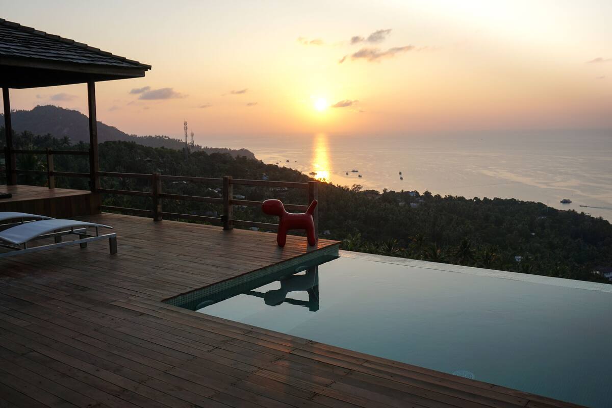 Una terraza de madera con piscina, una silla roja y vistas a la puesta de sol sobre el mar.