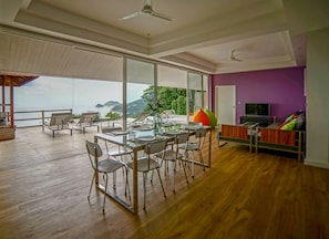 A spacious living area with a dining table set for four, surrounded by chairs, a sofa, and a television. Large windows offer a view of the outdoors. - Naroua Villas (Koh Tao)
