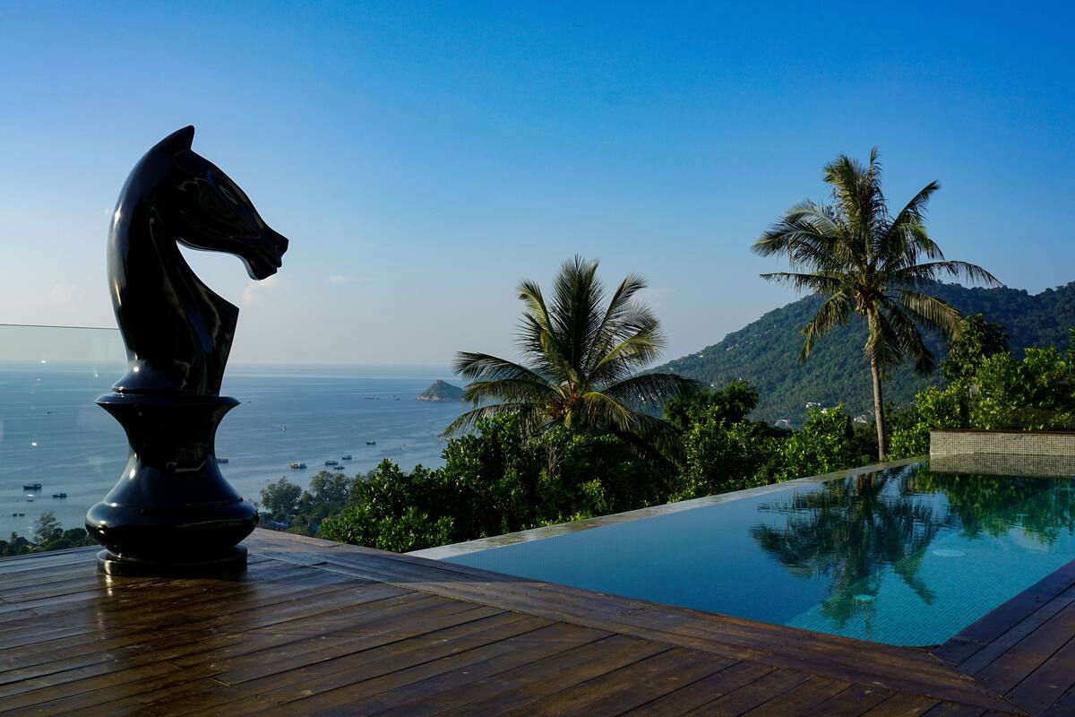 Una terraza de madera con una escultura de un caballo negro, una piscina y vistas al mar y a las montañas.
