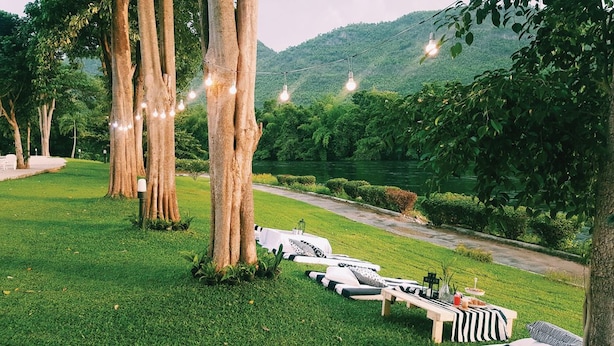 A serene outdoor setting with lounge chairs and a table set up under large trees, overlooking a river and lush greenery. - Aekpailin River Kwai Resort Kanchanaburi (Kanchanaburi)
