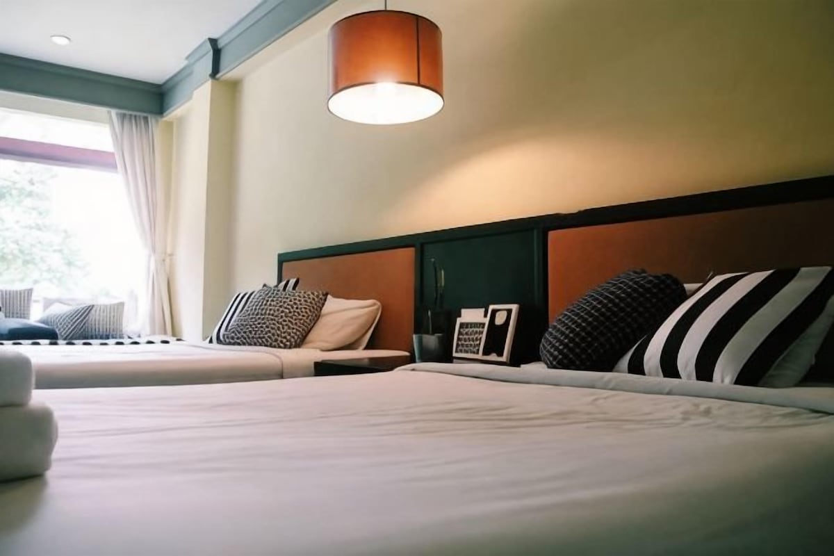 A hotel room with two beds, a headboard, a hanging light fixture, and a window with curtains.