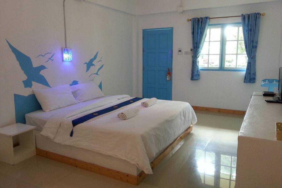 Ein Schlafzimmer mit Bett, Nachttisch, Schreibtisch mit Computer, Fenster mit Vorhängen und einer blauen Tür.