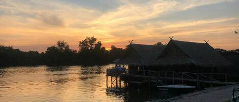 Vista al atardecer con chozas de techo de paja a orillas de un río tranquilo.