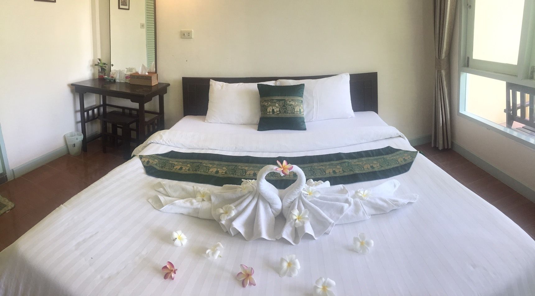 A neatly made bed with a decorative towel arrangement and flowers, a small table with a mirror, and a window with curtains.