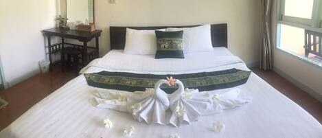 A neatly made bed with a decorative towel arrangement and flowers, a small table with a mirror, and a window with curtains.