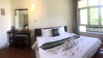 A hotel room with a bed, a desk, a chair, a mirror, and a window with a view of greenery.