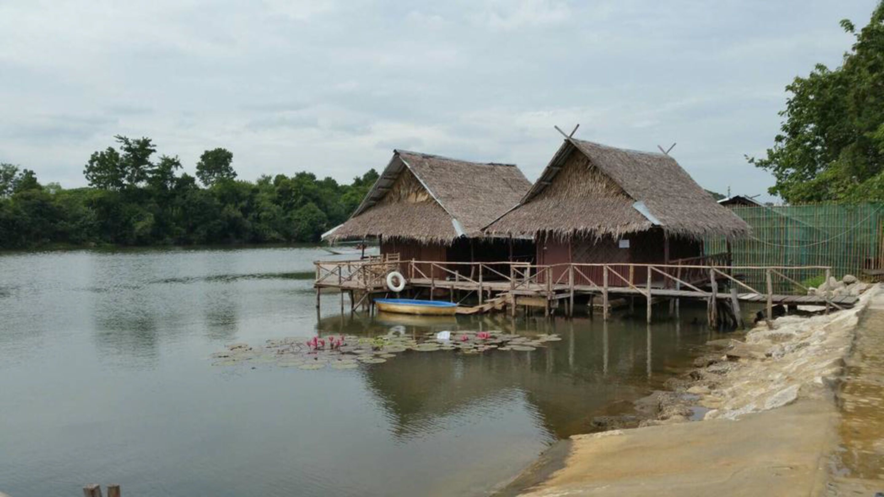 A thatched-roof structure with a wooden deck extending over water, surrounded by greenery.