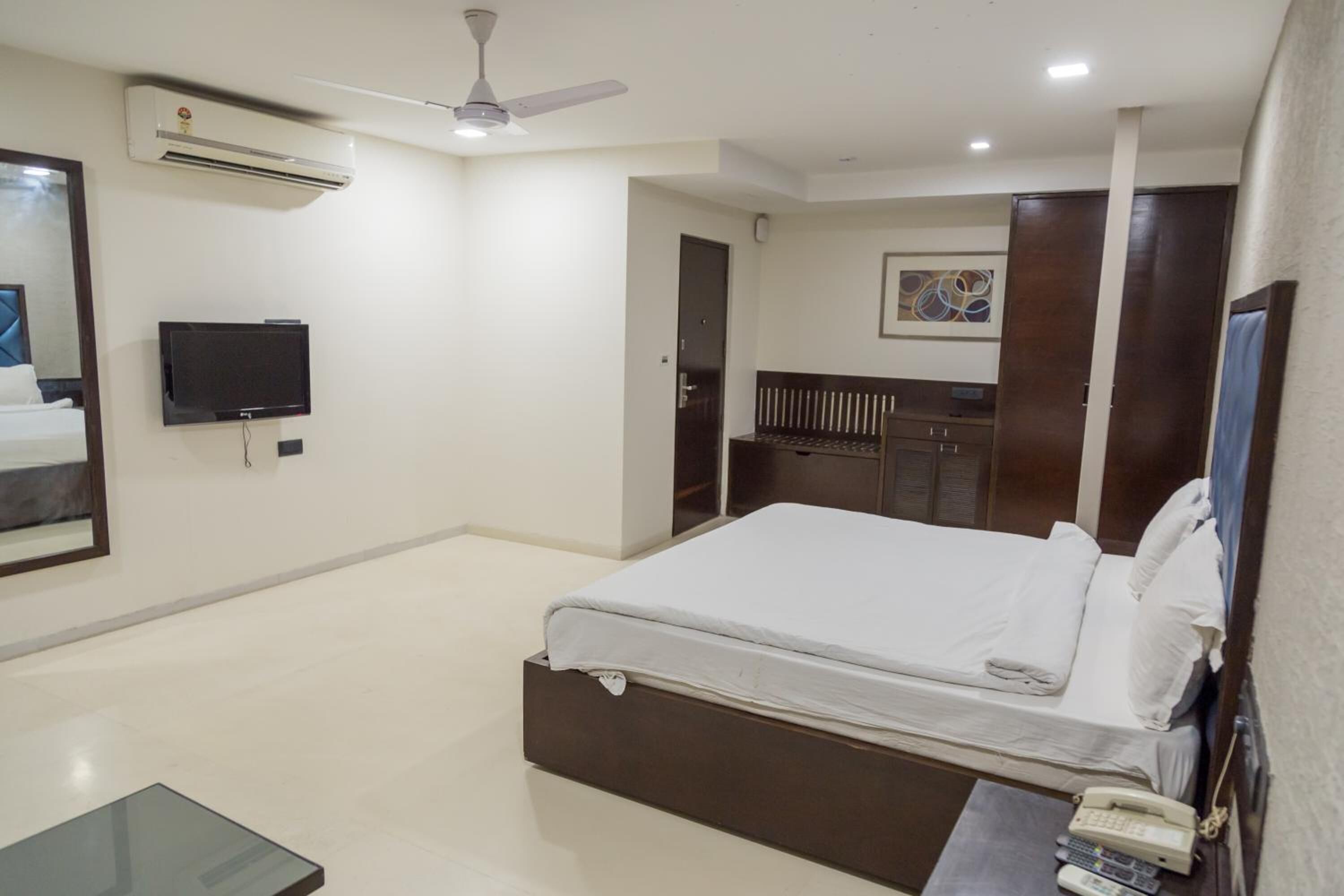 A hotel room with a bed, a TV, an air conditioner, a ceiling fan, and a desk with a phone.
