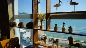 Une vue à travers une fenêtre, avec des étagères où sont exposés des objets décoratifs, un vase de fleurs et un paysage marin.