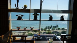 Une pièce dotée d’une grande fenêtre donnant sur une plage, avec une table en bois sur laquelle se trouvent une théière, des tasses et une bouteille. Des étagères au-dessus de la table sont ornées de divers objets décoratifs.