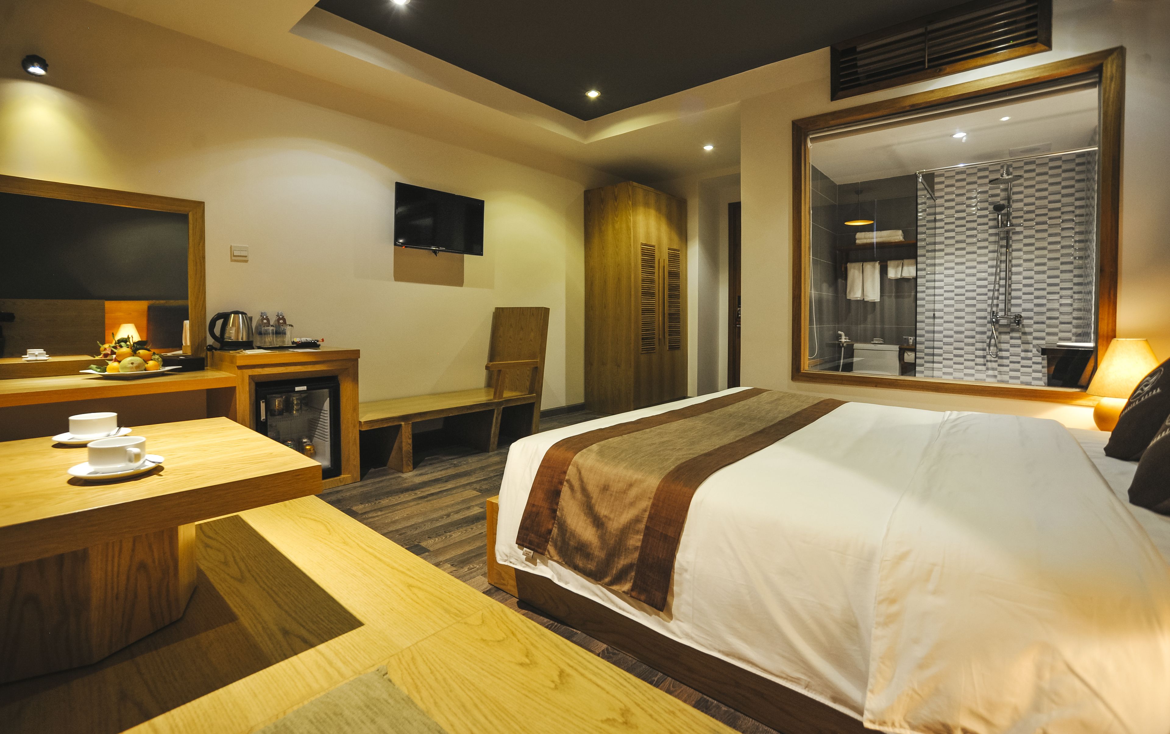 A hotel room with a bed, a desk with a chair, a television, and a bathroom visible through a large window.