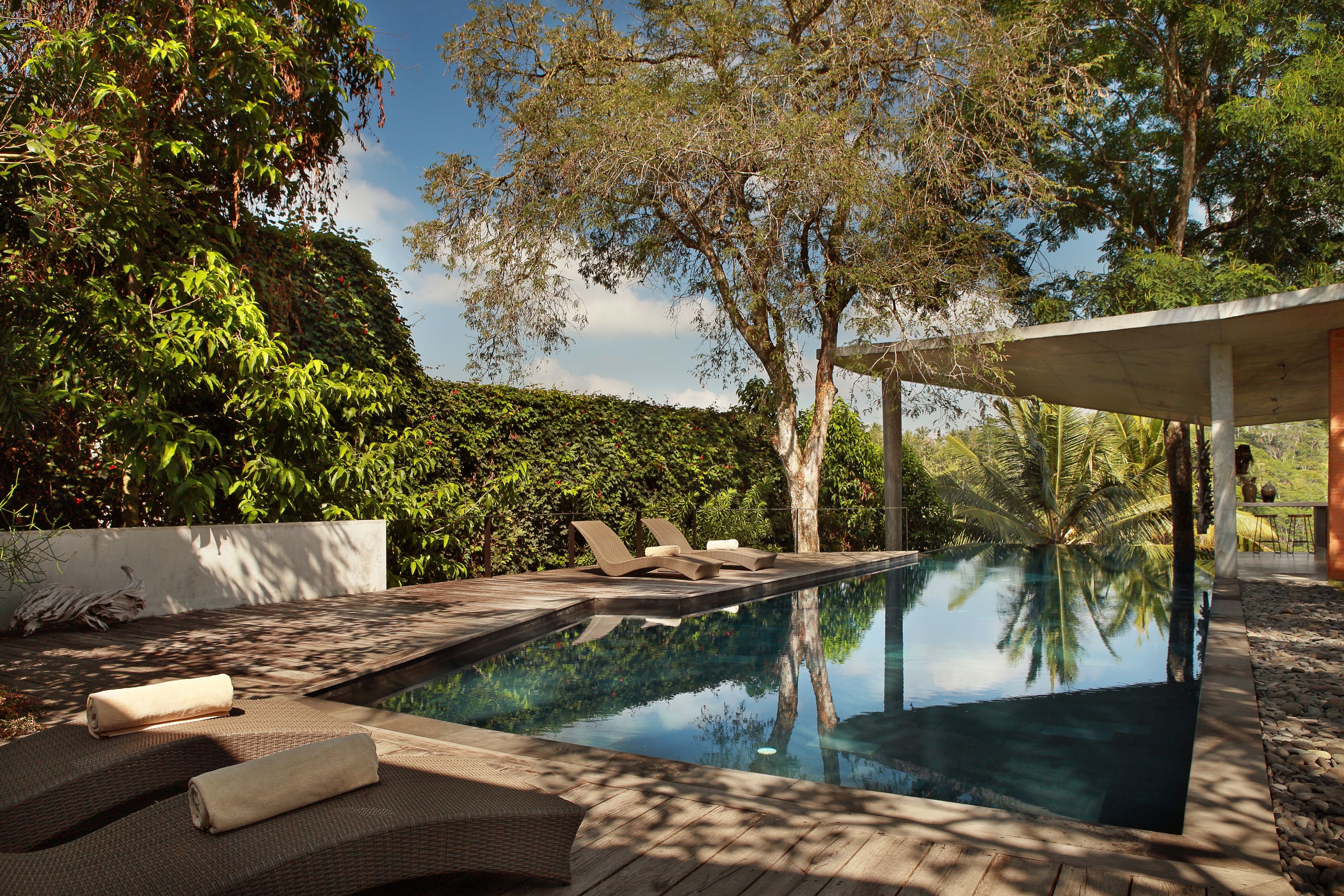An infinity pool with lounge chairs and a wooden deck, surrounded by lush greenery and trees.